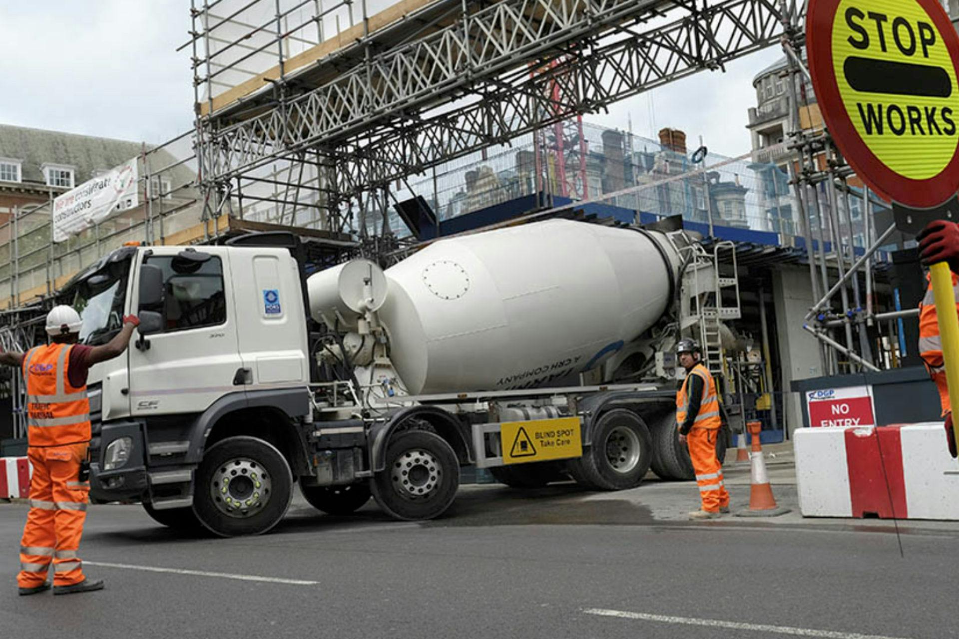 Lorry exiting a building site with workmen guiding them out and stopping traffic